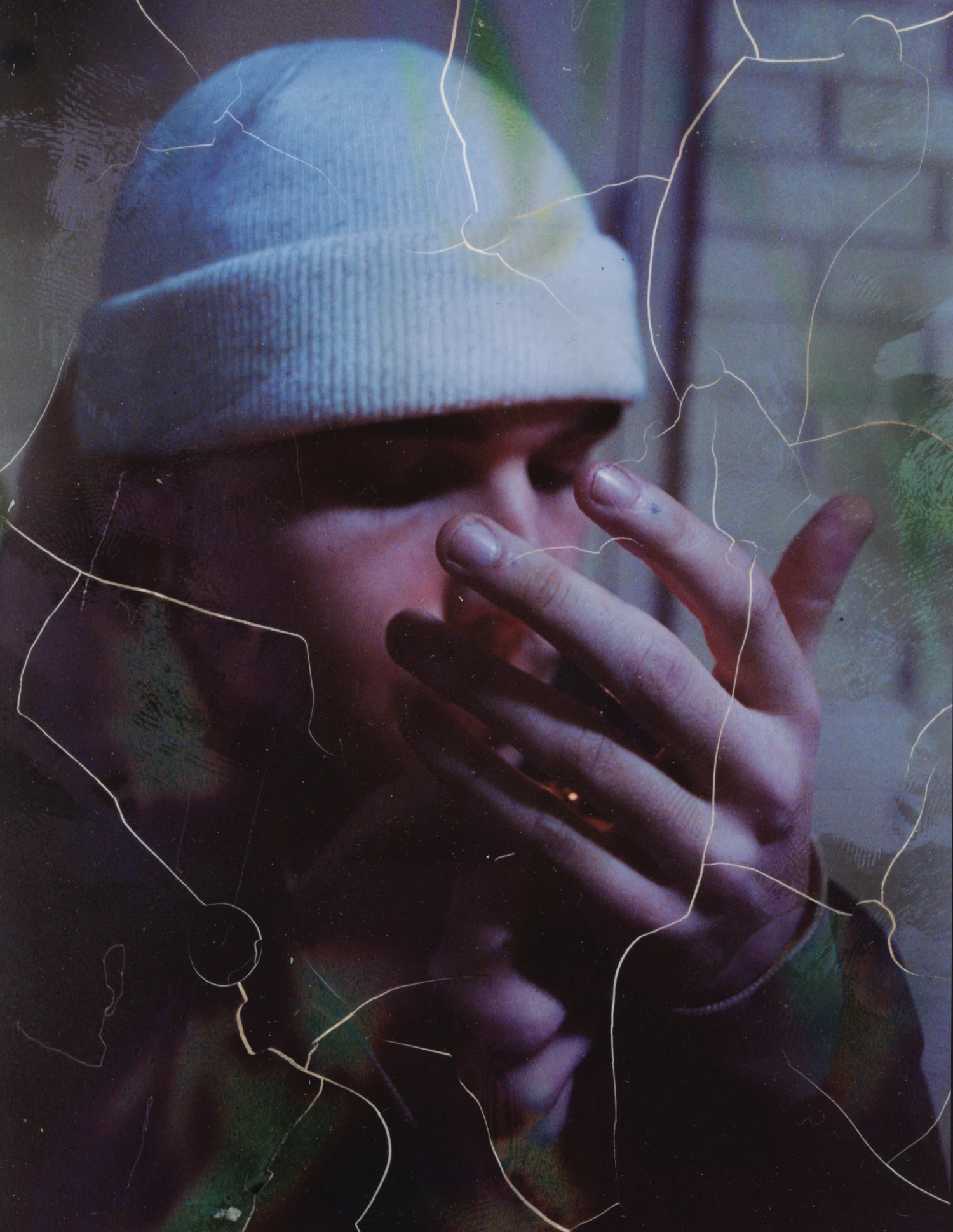 A grainy portrait of a person wearing a knit cap with their hands over their face