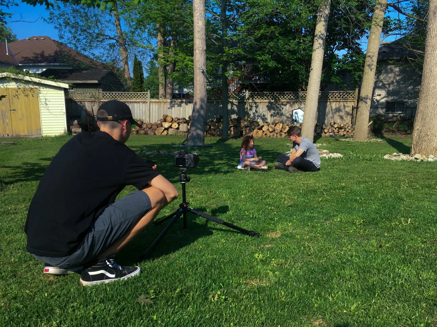 A camera operator filming two people sitting on a backyard lawn