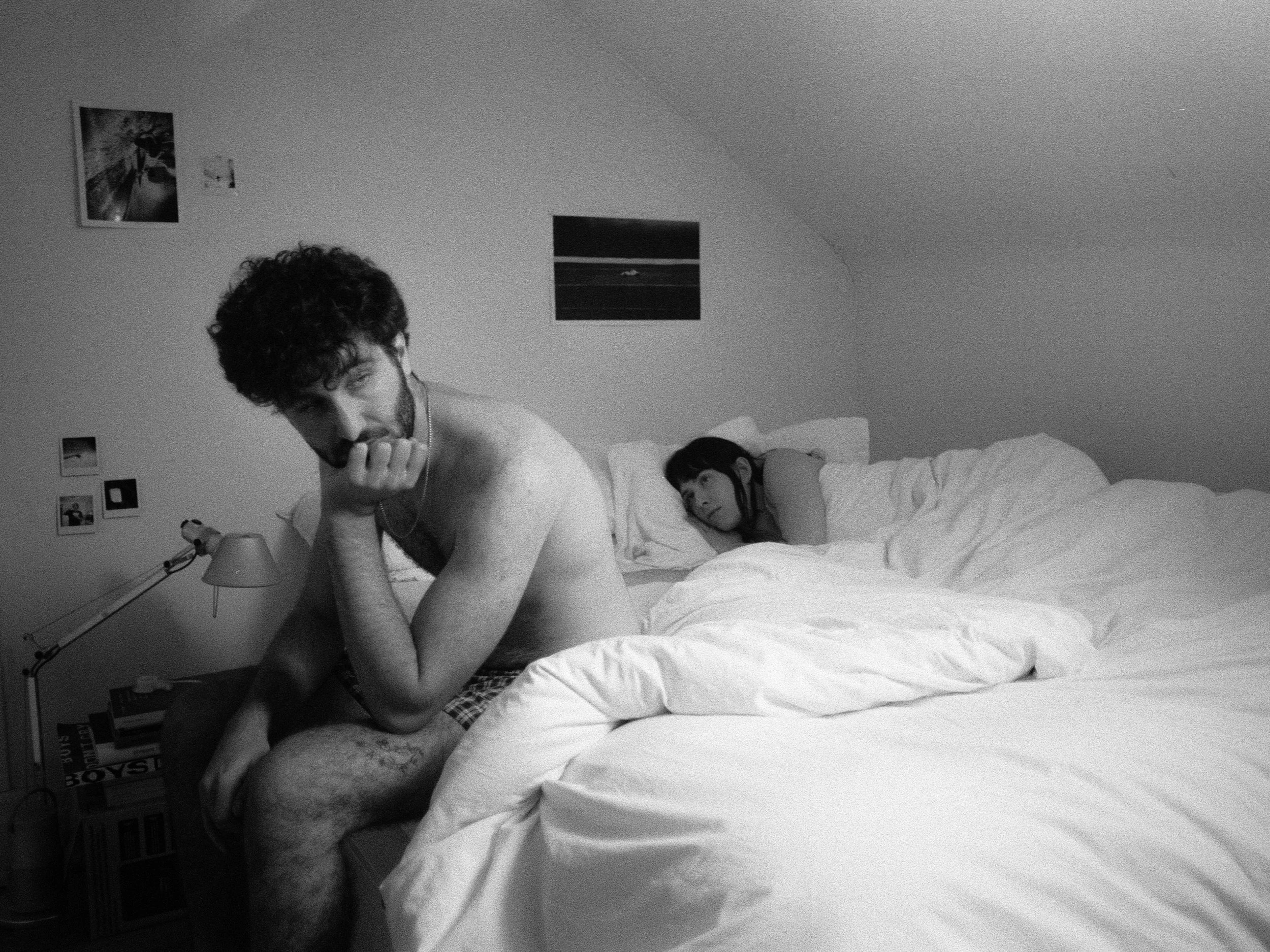Black-and-white film still of a man sitting on the edge of a bed while a woman lies behind him