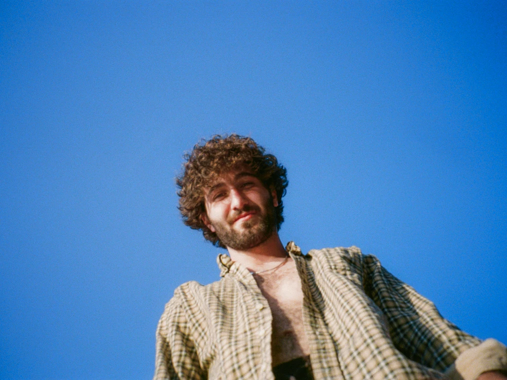 A curly-haired man in an open shirt against a clear blue sky